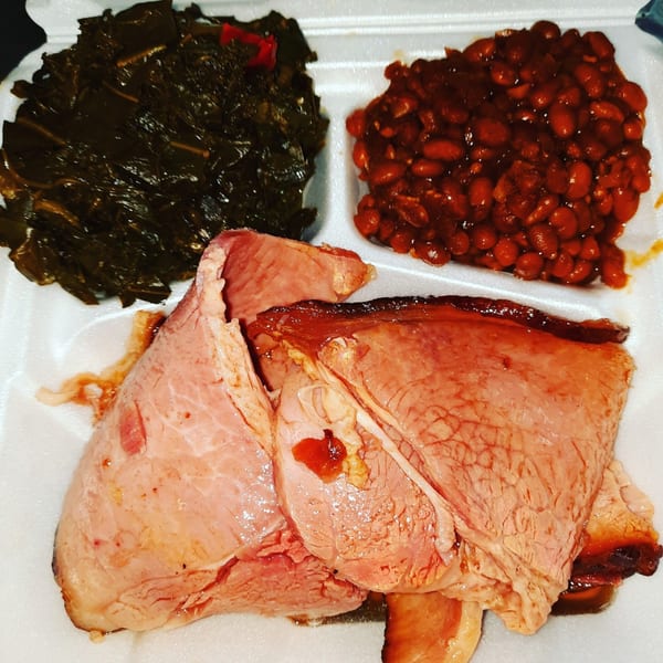Village BBQ's smoked meat with collard greens and baked beans in a white takeout container.