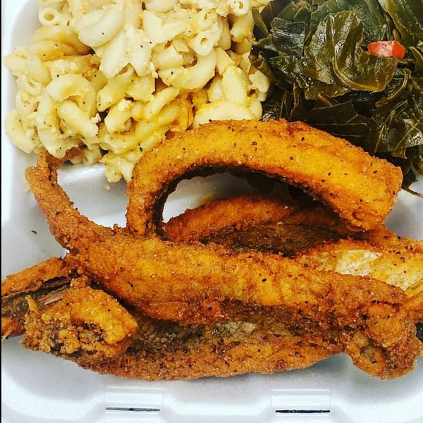 Fried fish, macaroni and cheese, and collard greens in a white takeout container.